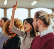 A Caucasian businesswoman is holding her hand up in a conference room to ask the speaker a question.