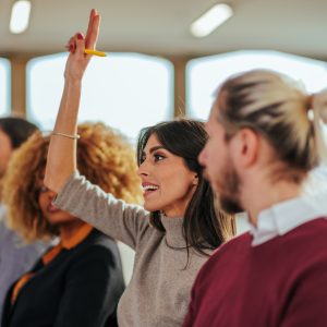 A Caucasian businesswoman is holding her hand up in a conference room to ask the speaker a question.