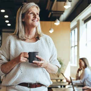 Thoughtful businesswoman standing in a co-working space. Mature businesswoman smiling cheerfully while holding a cup of coffee. Experienced entrepreneur contemplating new ideas during her coffee break.